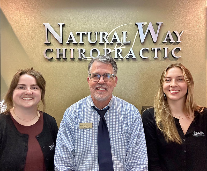 everett-june-nine Three smiling people pose in front of a Natural Way Chiropractic sign inside the clinic.