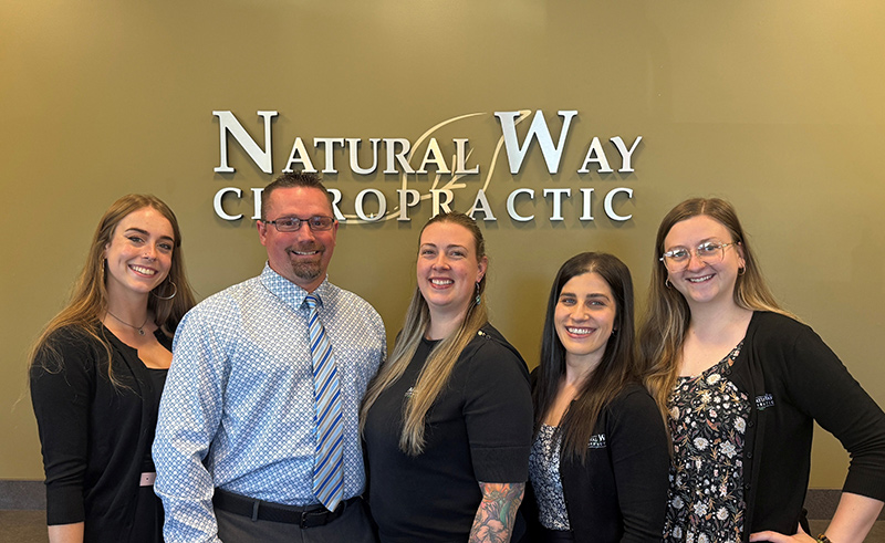 fern-june Five staff members stand together and smile for a photo in front of a Natural Way Chiropractic sign on a warm olive wall, representing a friendly clinic team.