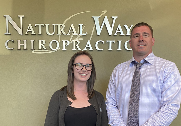 van-staff Two staff members pose for a photo in front of a Natural Way Chiropractic sign on a green wall.
